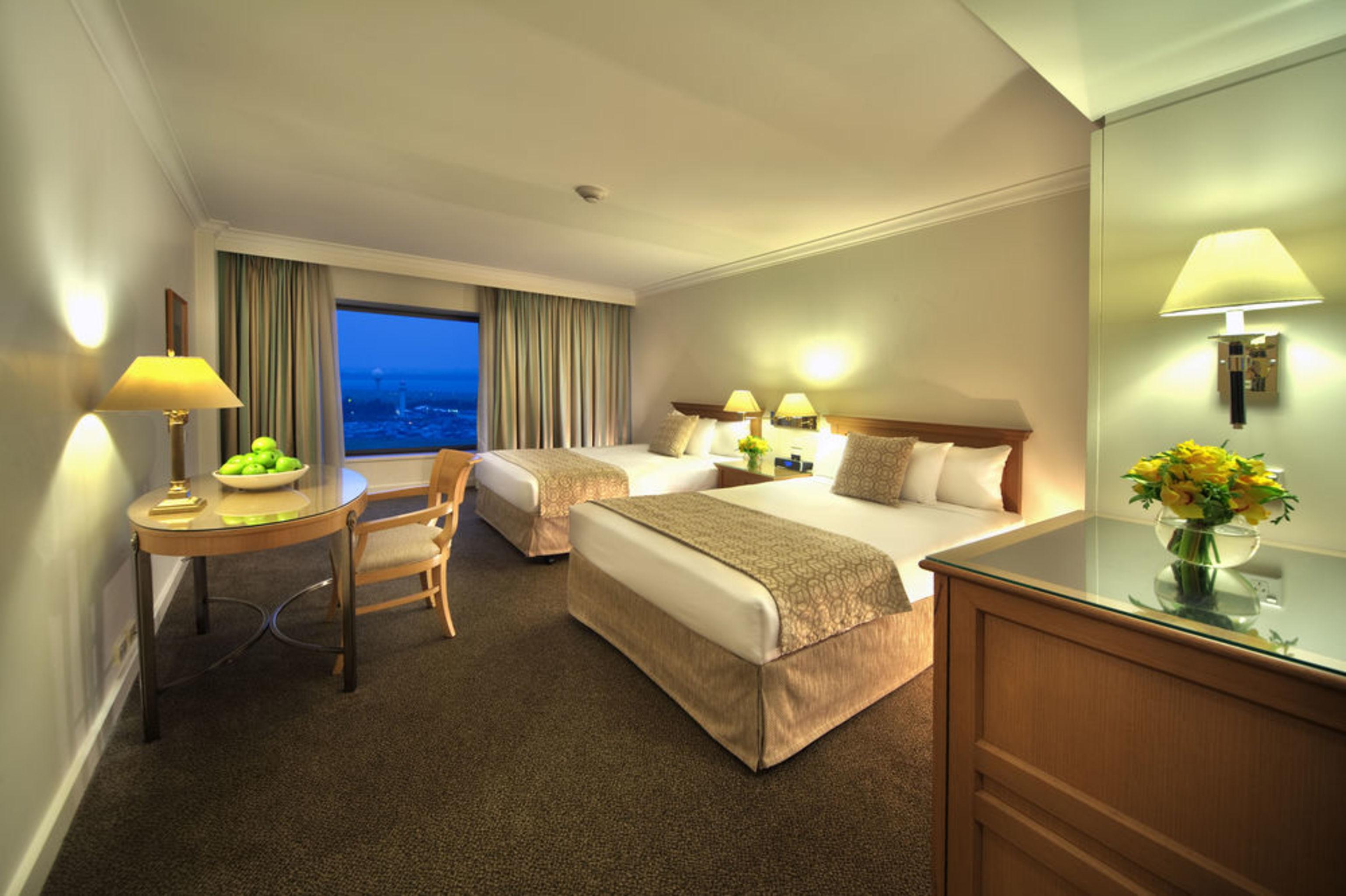 Stamford Plaza Sydney Airport Hotel & Conference Centre Room photo The photo shows a hotel room featuring two large beds with light-colored linens and decorative pillows. There is a wooden table with a chair placed nearby, and a bowl of fruit is on the table. The room is well-lit with lamps on either side of the bed