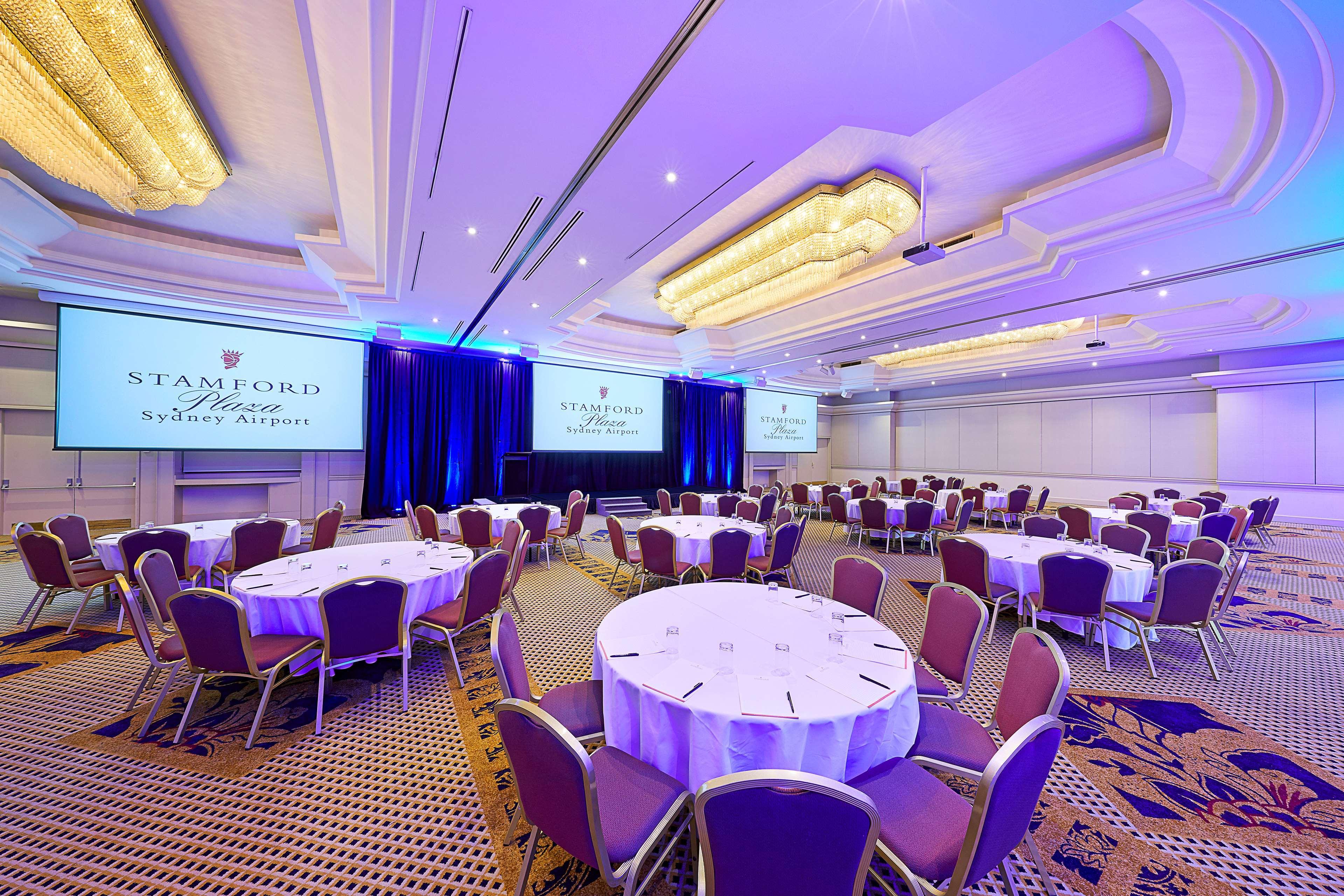 Stamford Plaza Sydney Airport Hotel & Conference Centre Exterior photo The photo depicts an elegantly designed event space, likely set up for a conference or banquet. It features round tables, each with white tablecloths, arranged in a spacious layout. The chairs surrounding the tables are upholstered in purple fabric. 