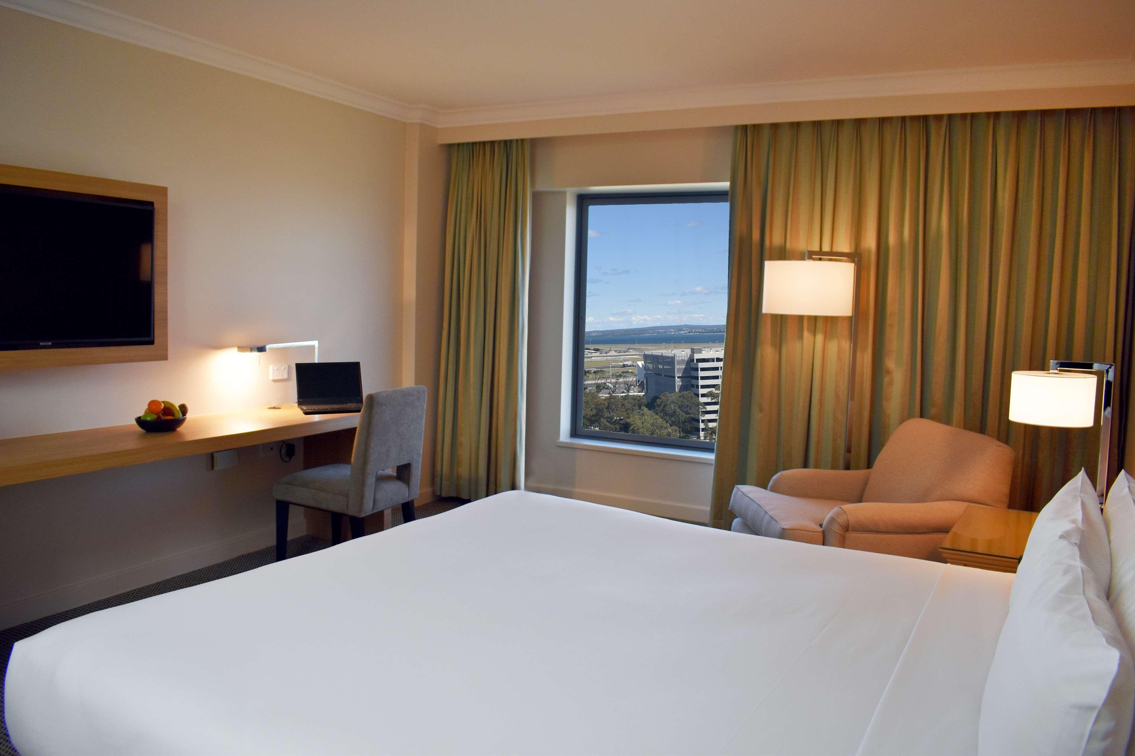 Stamford Plaza Sydney Airport Hotel & Conference Centre Exterior photo The photo shows a hotel room with a modern and clean design. There is a large bed with white bedding in the foreground. To the side, there is a small seating area with an armchair and a lamp. A sleek desk with a chair is positioned against the wall n