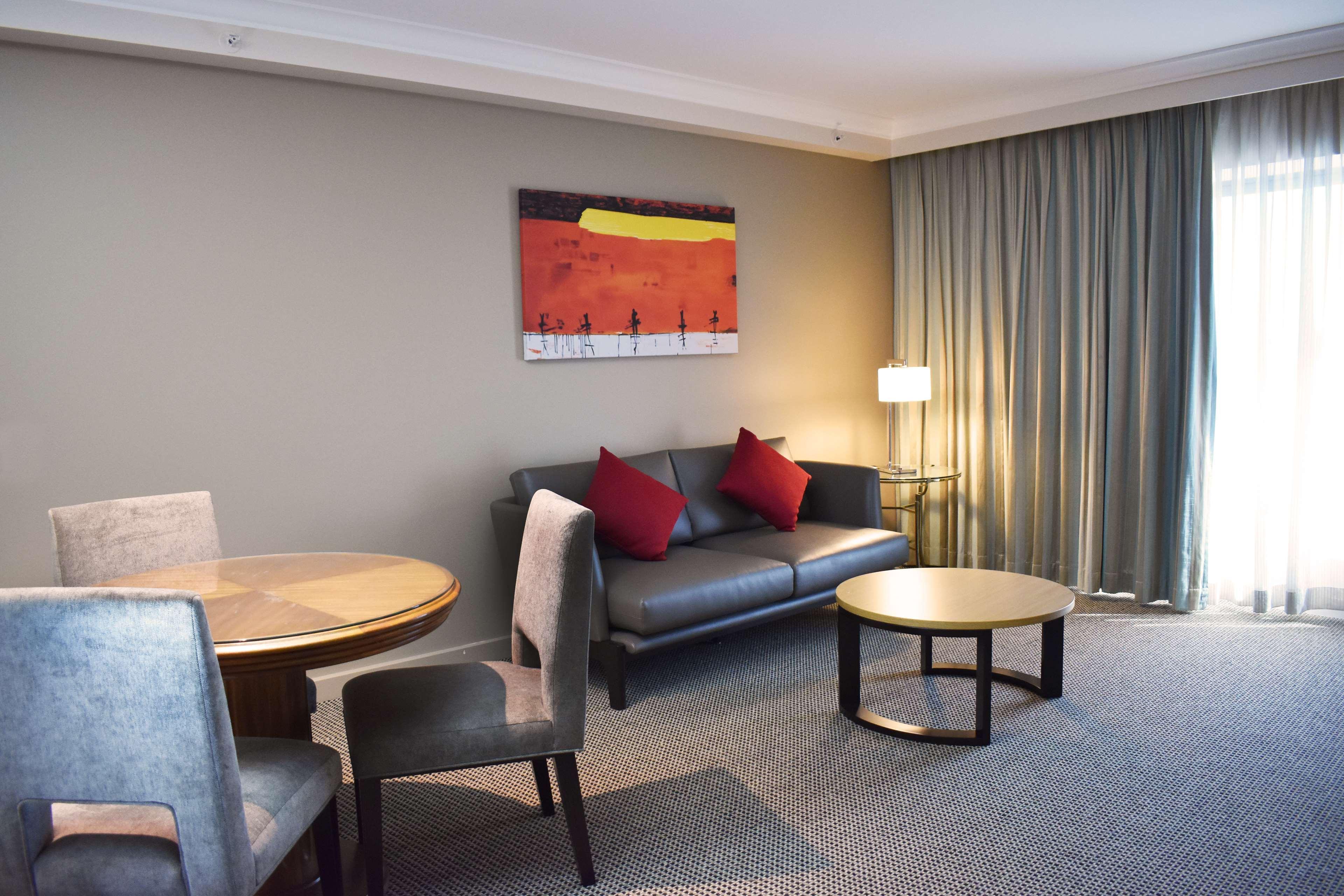 Stamford Plaza Sydney Airport Hotel & Conference Centre Exterior photo The photo shows a modern living area in a hotel suite. There is a gray sofa with red throw pillows, and a round coffee table in front of it. To the side, there is a small dining table with chairs. The walls are painted a light color, and there is a l