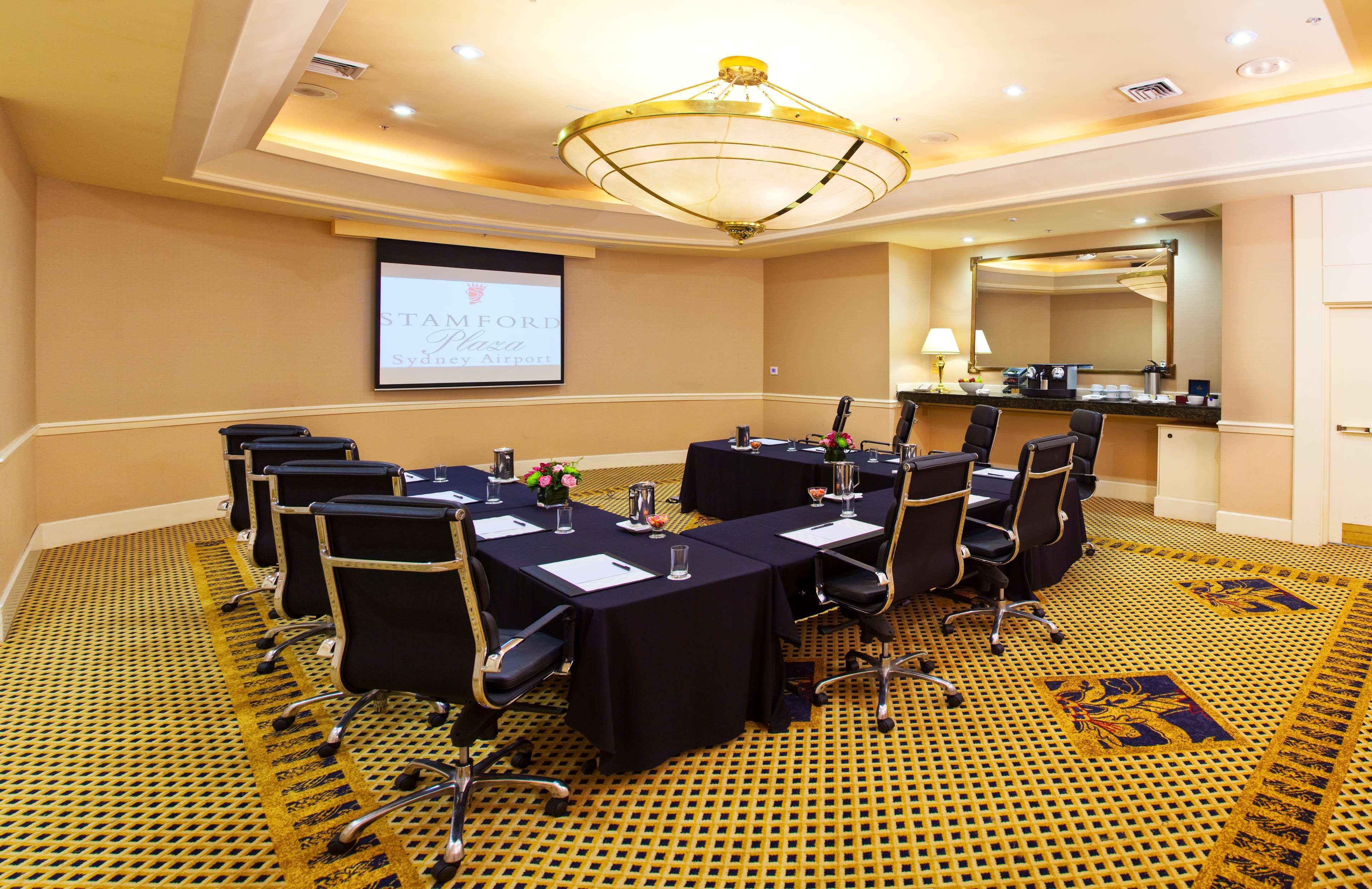 Stamford Plaza Sydney Airport Hotel & Conference Centre Exterior photo The photo shows a conference room set up for a meeting. In the center, there is a rectangular table covered with a black tablecloth, surrounded by high-backed chairs. Each chair has a notepad and pen placed on the table in front of it. 

At the front
