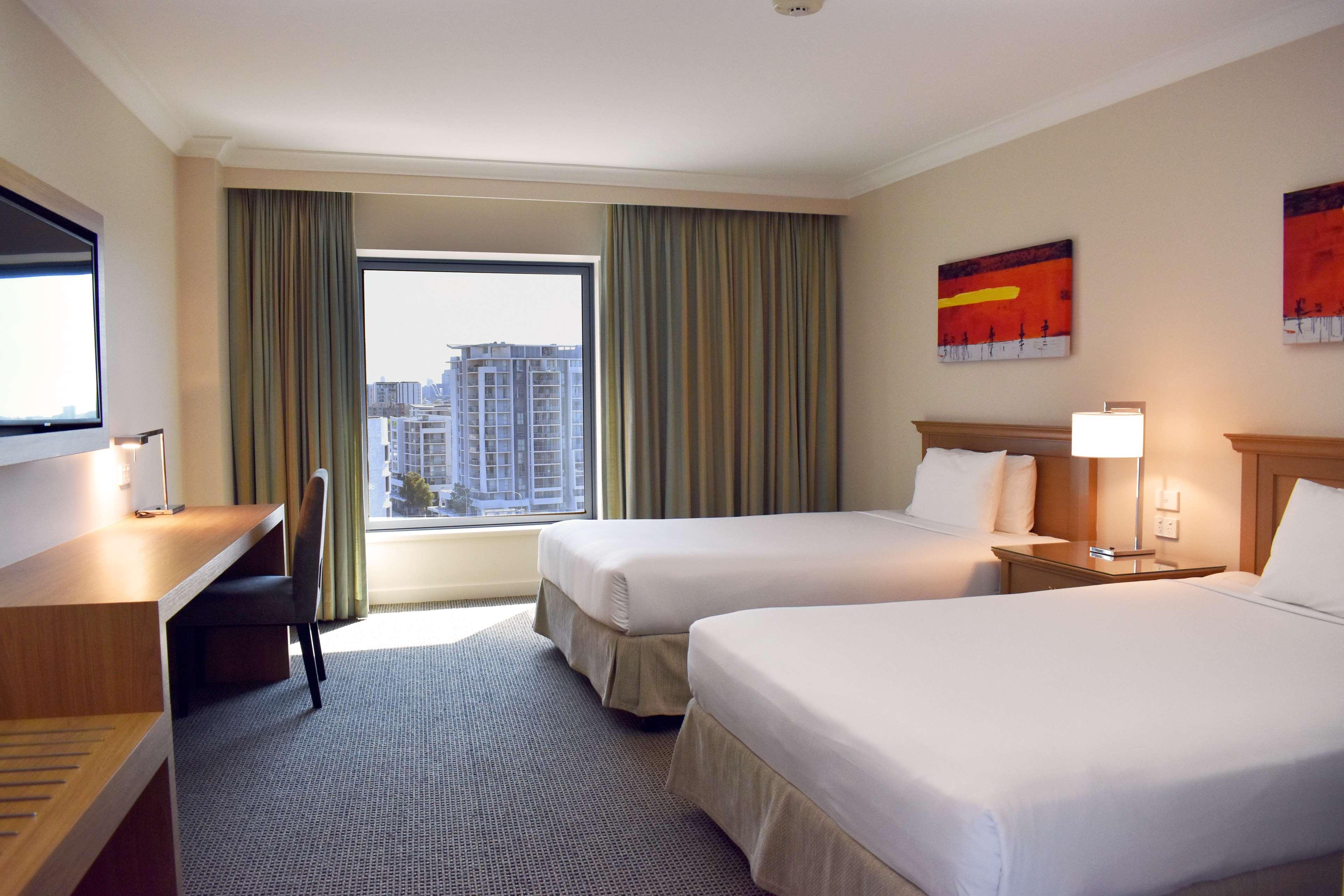 Stamford Plaza Sydney Airport Hotel & Conference Centre Exterior photo The photo shows a hotel room that features two single beds with white beddings. There is a desk and chair situated beside the window, which is dressed with light curtains. The room has neutral-colored walls and a carpeted floor. A piece of abstract a