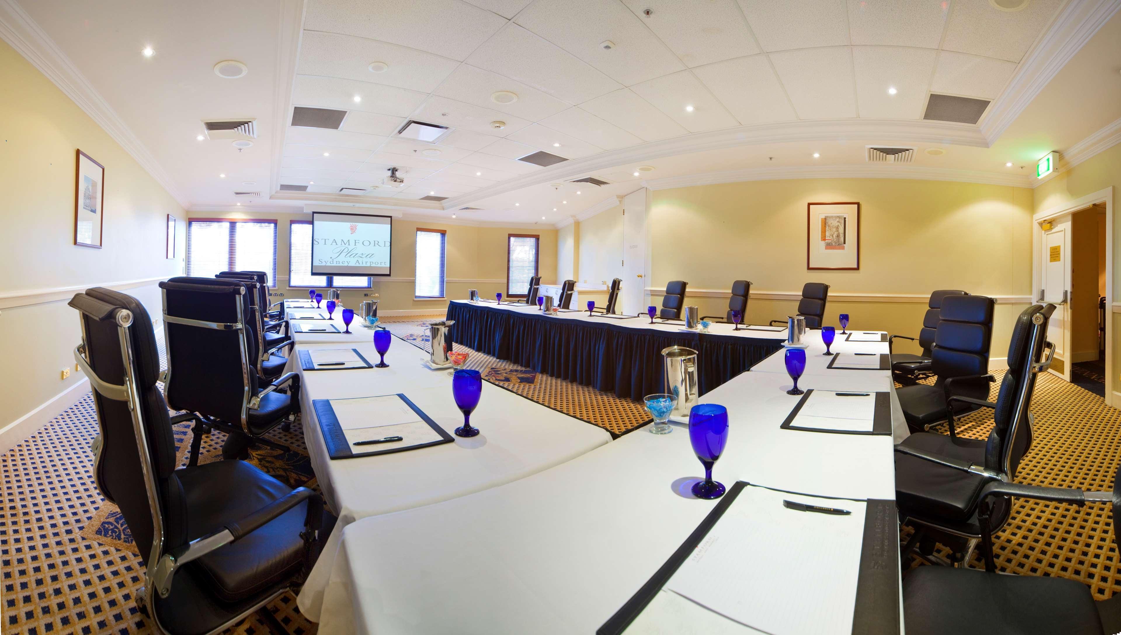 Stamford Plaza Sydney Airport Hotel & Conference Centre Exterior photo The photo shows a conference room set up for a meeting. There are several tables arranged in a U-shape, covered with white tablecloths. Each table is equipped with a notepad and a blue glass beside it. The room features several black chairs and has a