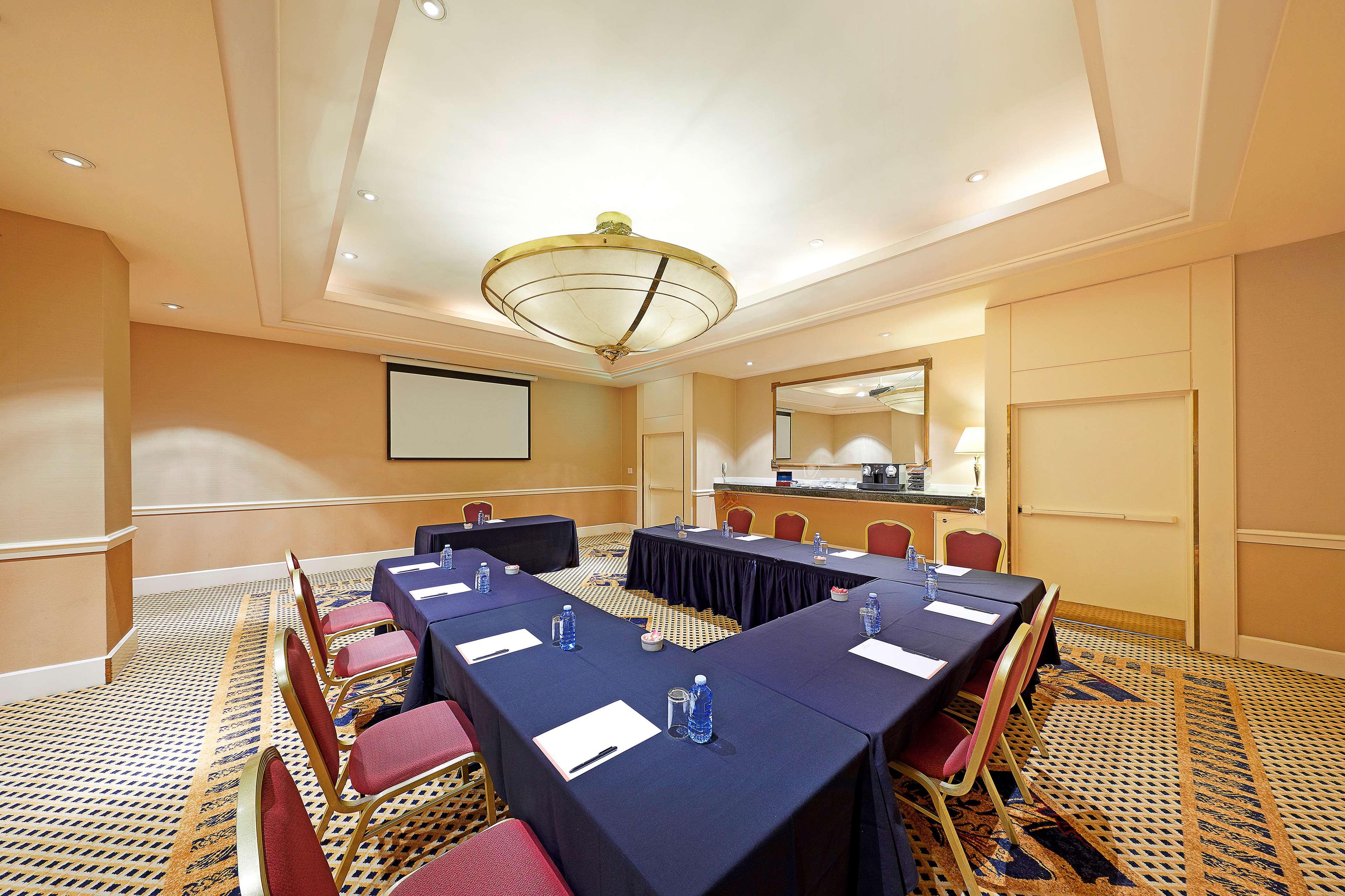 Stamford Plaza Sydney Airport Hotel & Conference Centre Exterior photo The photo shows a conference room set up for a meeting. There are two long tables arranged in a U-shape, covered with dark blue tablecloths. Each table has a few pieces of paper and a pen placed on top. The room features a chandelier hanging from the