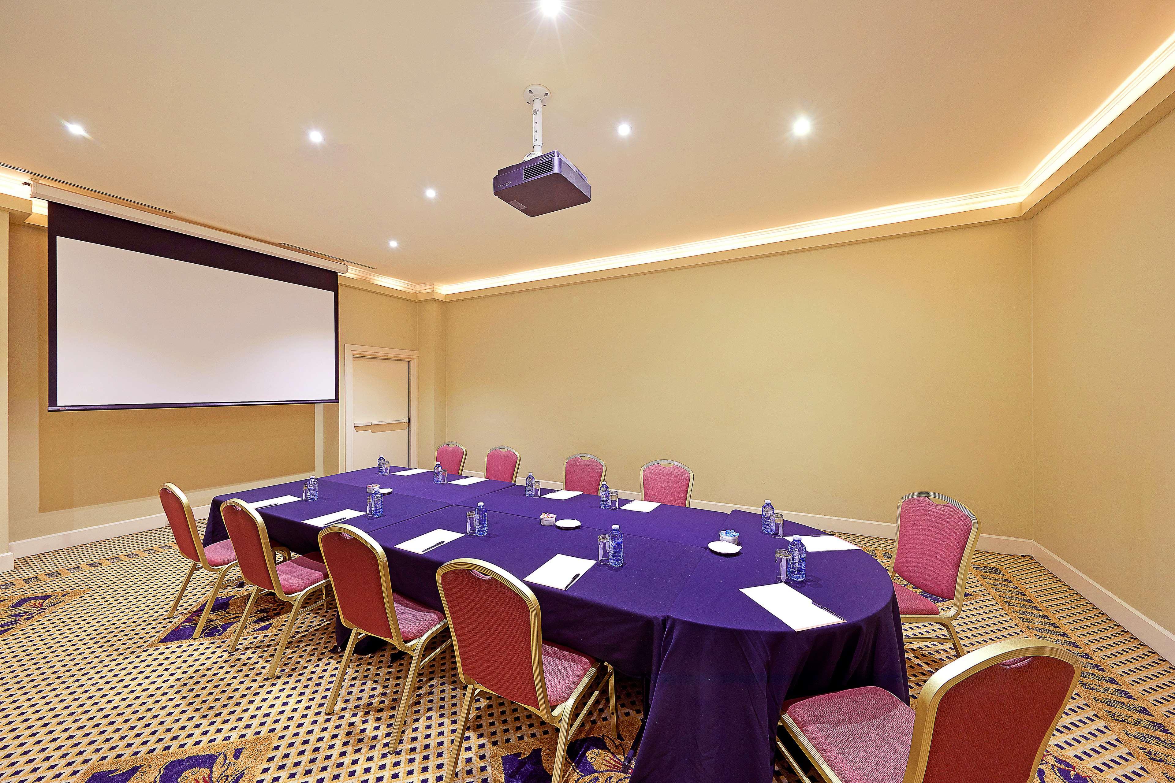 Stamford Plaza Sydney Airport Hotel & Conference Centre Exterior photo The photo shows a conference room setup. In the center, there is a round table covered with a dark purple tablecloth. Around the table, there are several pink chairs arranged neatly. On the table, there are some papers, cups, and water bottles. A pro