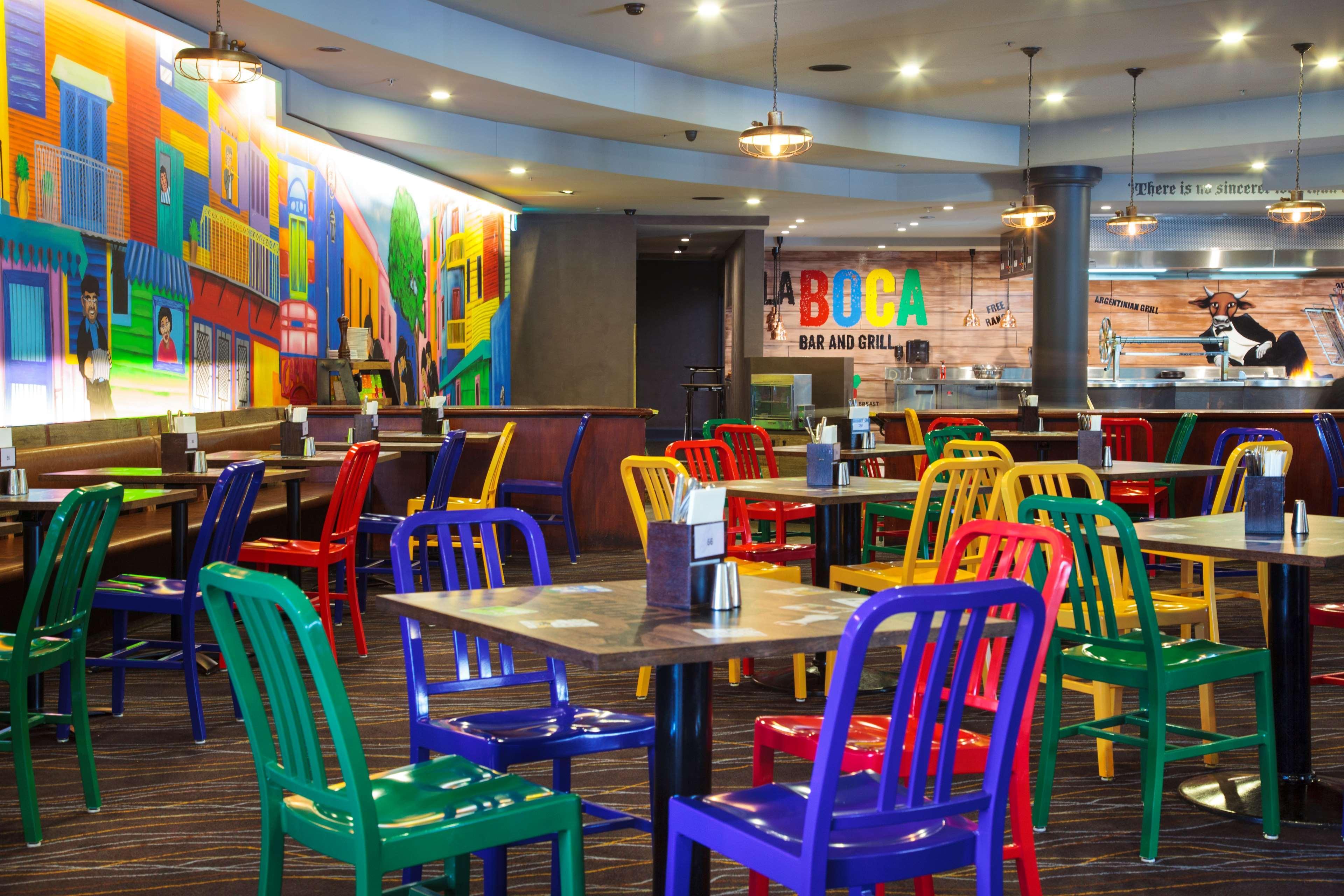 Stamford Plaza Sydney Airport Hotel & Conference Centre Exterior photo The photo shows the interior of a vibrant restaurant with a colorful decor. The seating area features tables surrounded by brightly colored chairs in various shades, including red, blue, green, and yellow. One wall has a large, colorful mural depicti