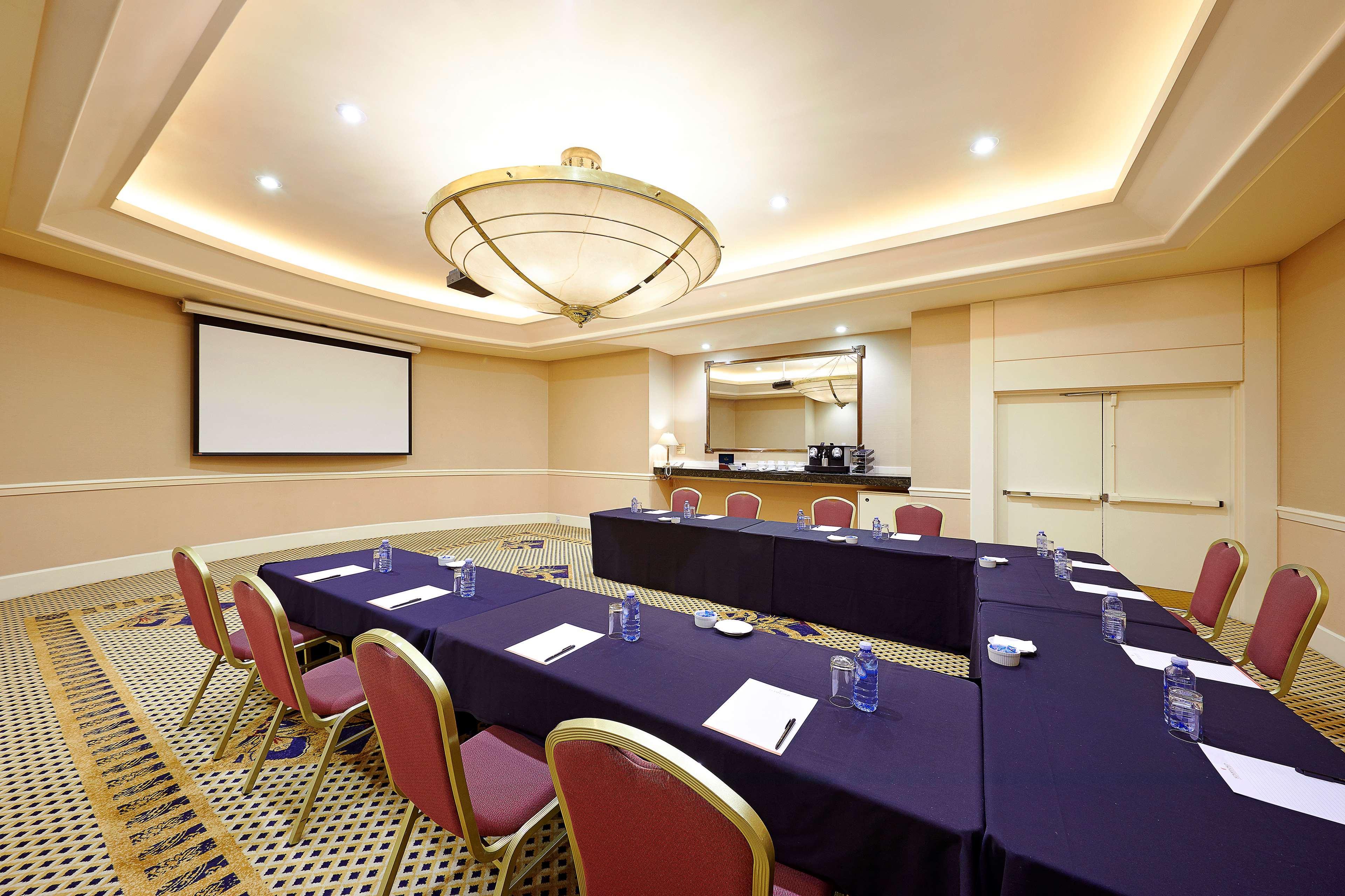 Stamford Plaza Sydney Airport Hotel & Conference Centre Exterior photo The photo shows a conference room setup. There are several rectangular tables arranged in a U-shape, covered with dark blue tablecloths. Each table has chairs with pink upholstery around it. On the tables, there are clear water glasses and paper note