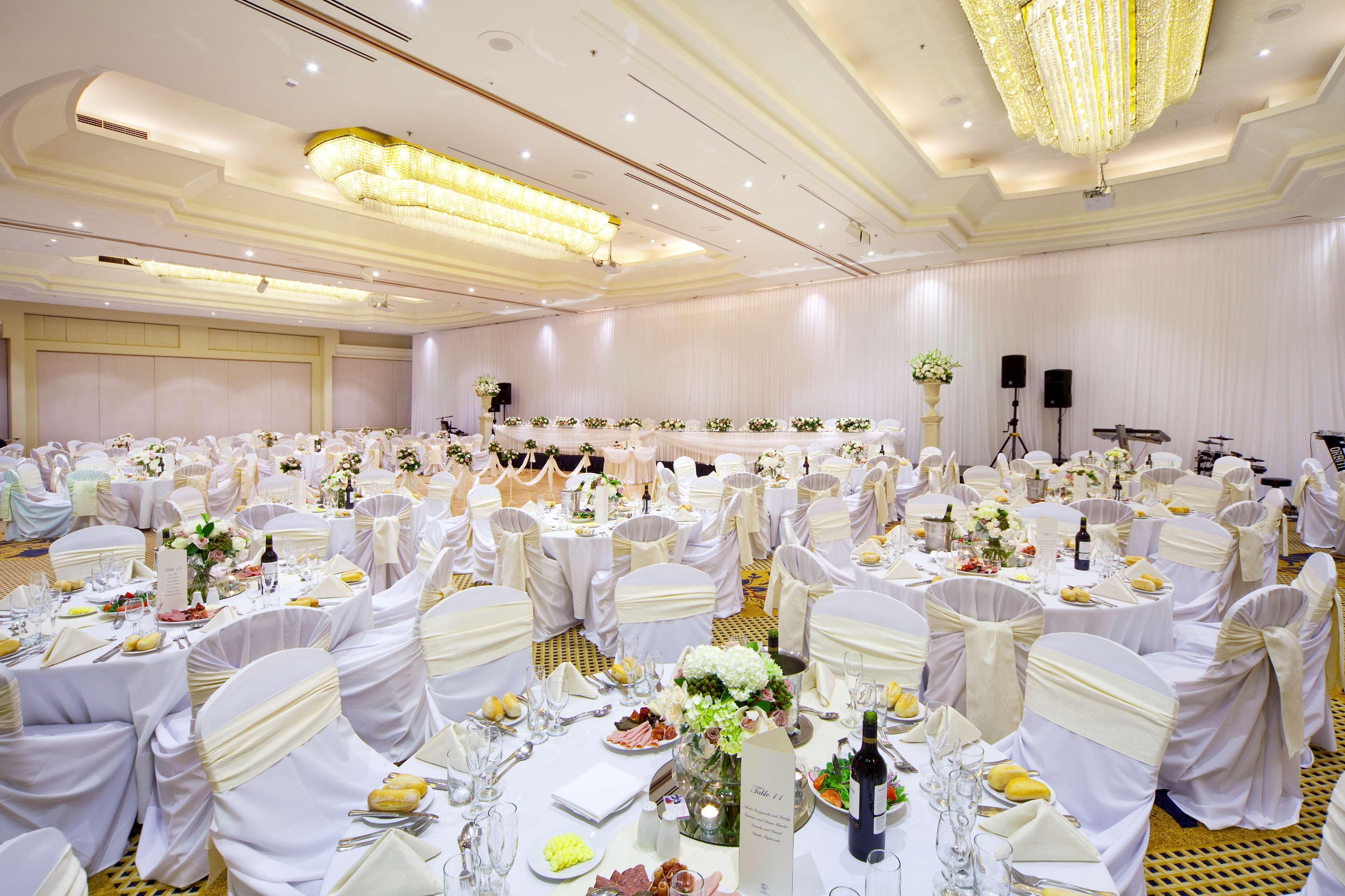 Stamford Plaza Sydney Airport Hotel & Conference Centre Exterior photo The photo shows a large banquet or event hall set up for a formal gathering. Several round tables are covered with white tablecloths and adorned with elegant decorations, including flower arrangements. Each table is set with plates, utensils, and gla