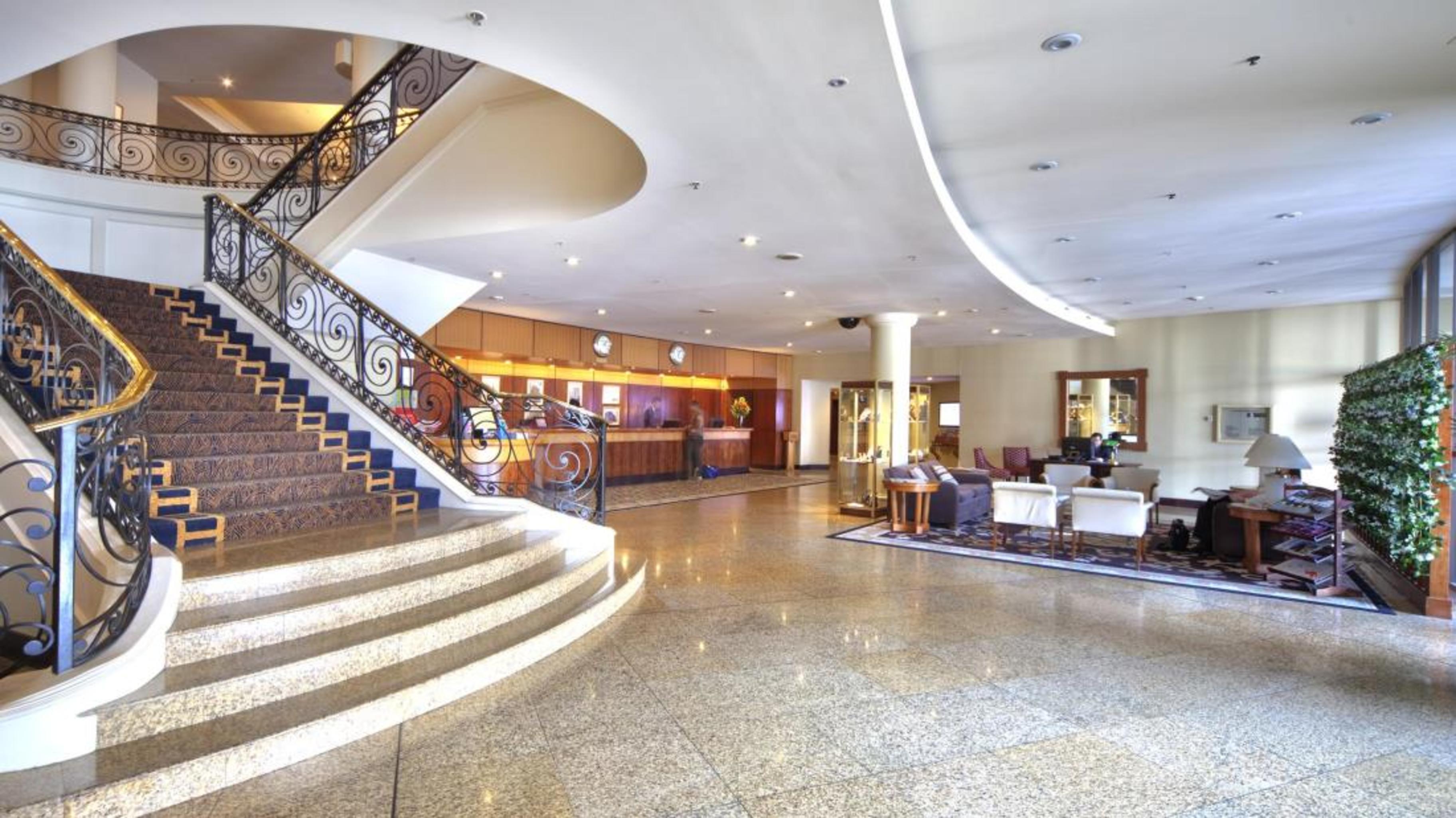 Stamford Plaza Sydney Airport Hotel & Conference Centre Exterior photo The photo shows the interior of a hotel lobby. There is a prominent staircase with elegant railings leading up to the upper level. The floor appears polished and shiny. To the left, there’s a reception area with a wooden desk where staff may assist g