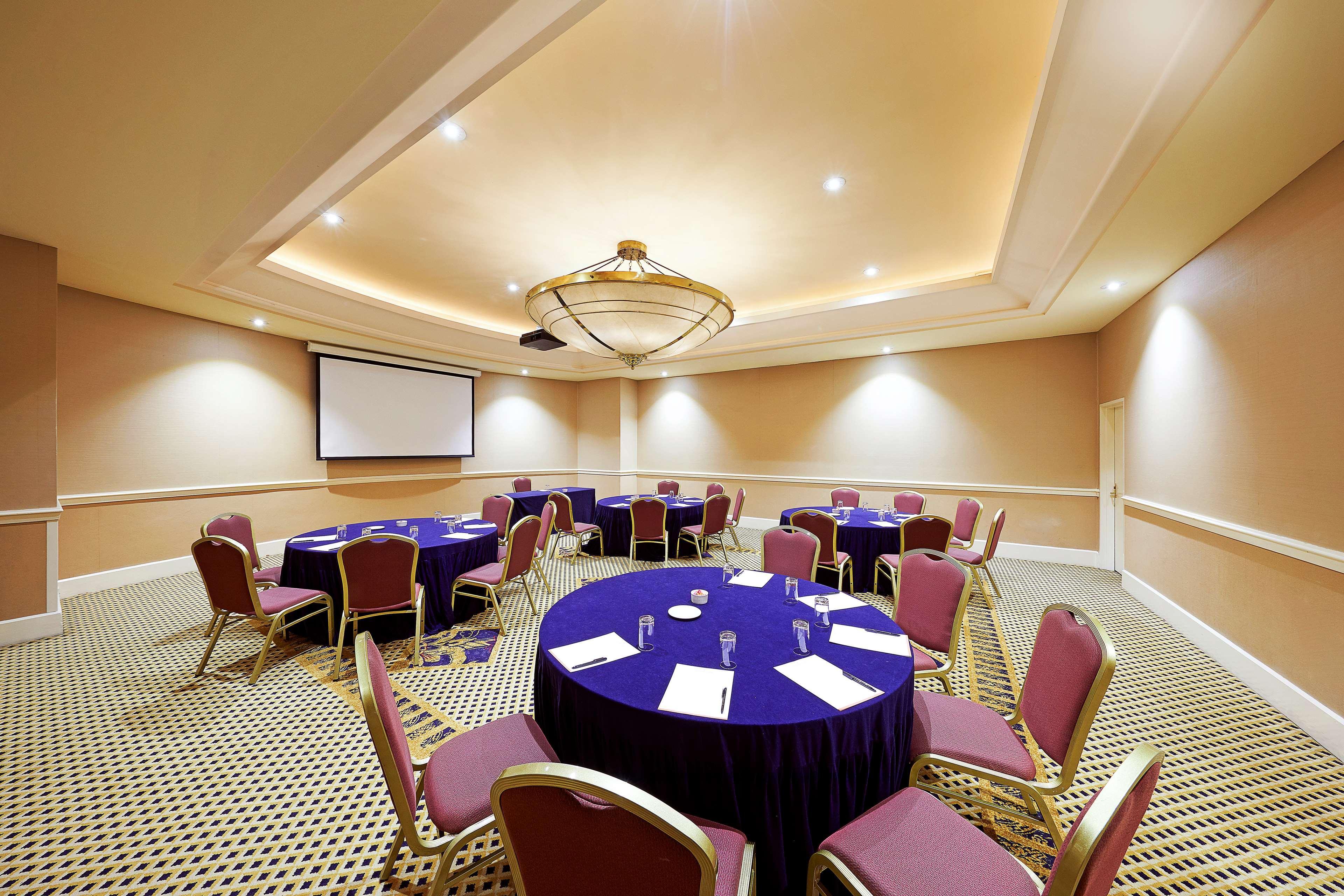 Stamford Plaza Sydney Airport Hotel & Conference Centre Exterior photo The photo shows a conference or meeting room. It features several round tables covered with purple tablecloths, each surrounded by pink chairs. There is a projector screen mounted on the wall, likely for presentations. The room has a warm lighting de