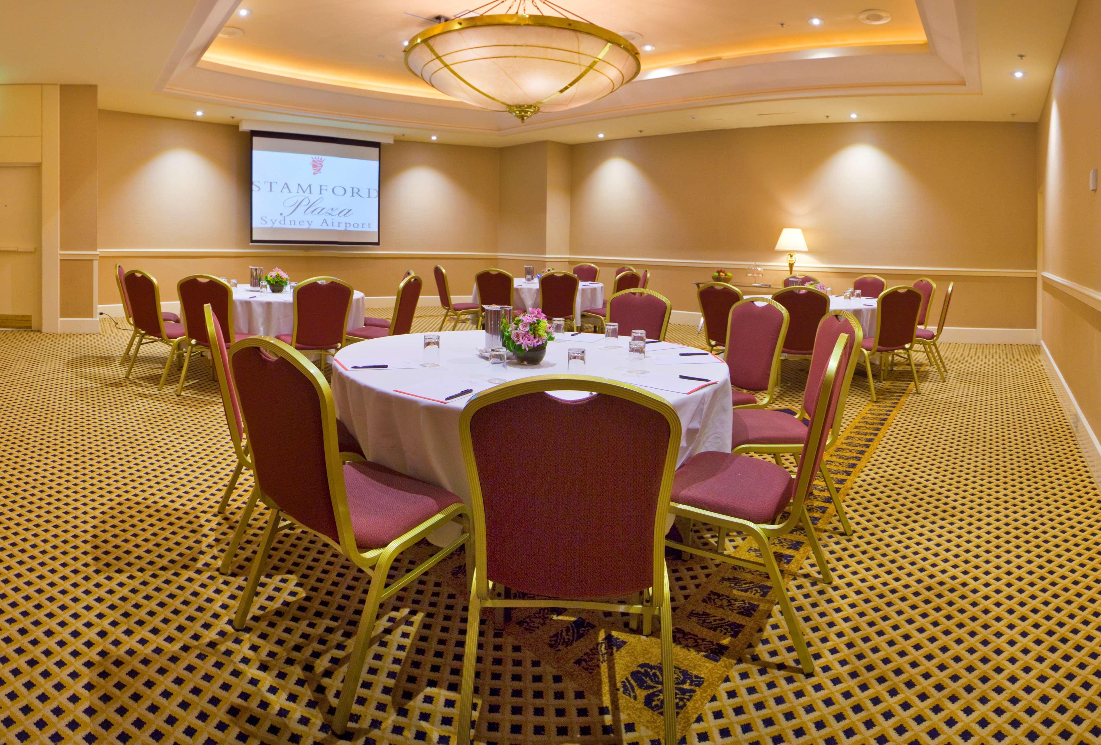 Stamford Plaza Sydney Airport Hotel & Conference Centre Business photo The photo shows a well-lit meeting or banquet room. In the center, there are several round tables covered with white tablecloths, each surrounded by elegant chairs with pink upholstery and gold frames. The tables are decorated with small floral arran